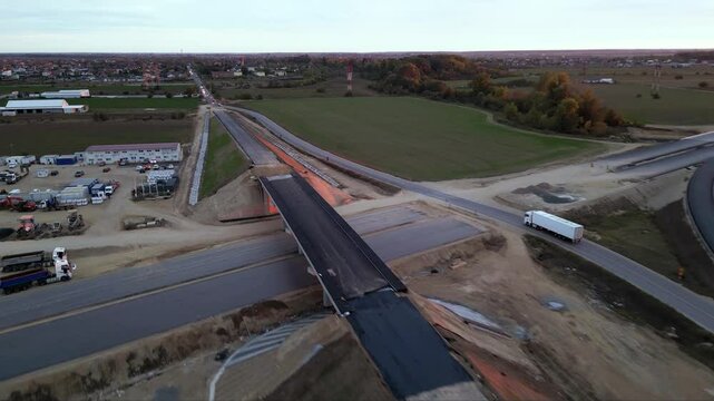 Low-altitude drone view from a newly paved highway overpass overlooking an active motorway during road construction. Modern transportation infrastructure with moving traffic, construction works and ex