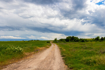 Empty Bulgarian country dirt road winding between green May fields under beautiful stormy sky in Haskovo Province, Southern Bulgaria