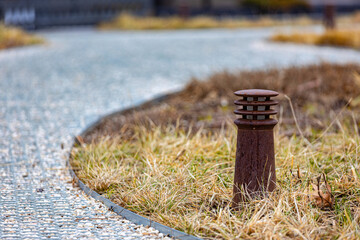 A weathered bollard stands beside a winding pebble pathway in a park, surrounded by dry autumn grass and a soft focus background.