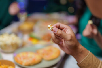 Hand holding a single piece of popcorn over a table laden with pizza, chips, and more popcorn, suggesting a casual party atmosphere
