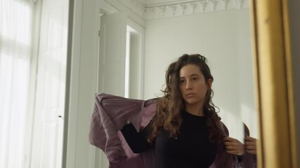 Young woman puts on a jacket or long sleeve shirt looking at herself in mirror. Confident female happy and feeling good about herself and body image. Smiling checking her fashion clothes in apartment