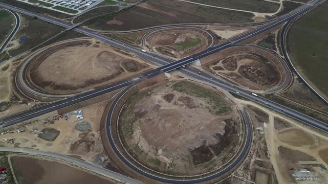 Top-down drone view of a newly built highway cloverleaf interchange with looping ramps, overpasses and active motorway traffic. Large-scale transportation infrastructure and civil engineering project 