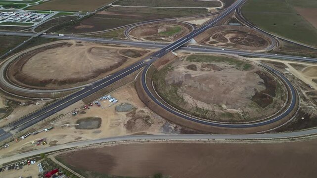 Top-down drone view of a newly built highway cloverleaf interchange with looping ramps, overpasses and active motorway traffic. Large-scale transportation infrastructure and civil engineering project 