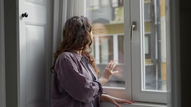 Sad unhappy woman stands by apartment window touching glass and looking outside. Female feeling lonely or depressed is waiting for someone or thinking and contemplating by city apartment window