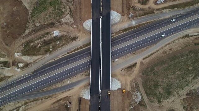 Top-down drone view of a newly built highway cloverleaf interchange with looping ramps, overpasses and active motorway traffic. Large-scale transportation infrastructure and civil engineering project 