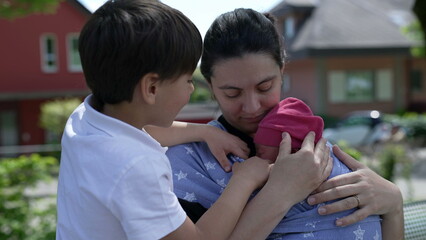 Mother holding newborn close in a sling with older son looking on, capturing a tender family...