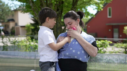 Mother holding newborn close on her chest in a sling while older son looks on attentively at park's...