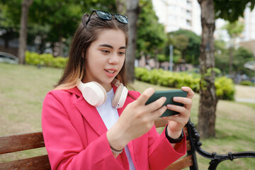 Young woman sitting on bench in park holding smartphone wearing headphones and pink jacket smiling and interacting with phone outdoors