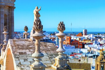 Jesus statue on top of Cathedral in Cadiz, Andalusia Spain. Sculpture is positioned on a rooftop, surrounded by ornate stone structures resembling flames. Panoramic city view with numerous buildings © Leilani