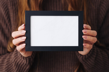 Woman holding empty photo frame. Blank space for advertising or poster. Black stroke of white surface. Stylish fingers with manicured nails. Promotional design. Woolen sweater clothing. Winter season.