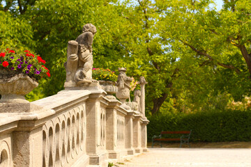 Eine elegante steinerne Balustrade mit kunstvollen Skulpturen und bl&uuml;henden Blumenk&uuml;beln vor einer gr&uuml;nen Parklandschaft. Perfekte Harmonie von Architektur und Natur in einem sommerlichen Garten