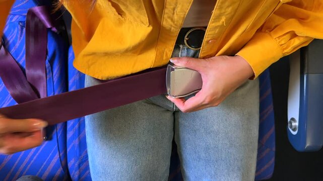 Closeup of an airplane Asian female passenger fastening a purple seatbelt while traveling flight. A woman wear bright yellow clothing contrasts with the deep purple belt, showcasing safety essentials.