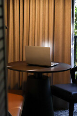 A laptop sits on a wooden table, its shadows stretching across the surface. A chair nearby complements the serene scene, with greenery softly framing the background