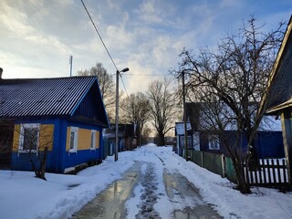 a road in the village in snow and with melted puddles, beautiful wooden houses on a sunny winter, fresh, cold day