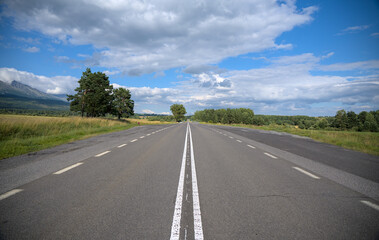 Fototapeta premium Straight road leading into the distance surrounded by fields and trees under a blue sky with clouds.