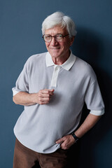 Vertical studio portrait of white haired senior man smiling at camera posing casually against blue...