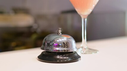 Gloved hand ringing a silver service bell on a bar counter with a pink cocktail in the background, highlighting hospitality and beverage service professionalism - Powered by Adobe
