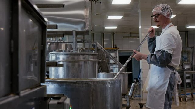 Medium full side shot of bearded Caucasian artisan male cheesemaker standing next to industrial metal vat, stirring milk mixture with cutting wire whisk during curdling process