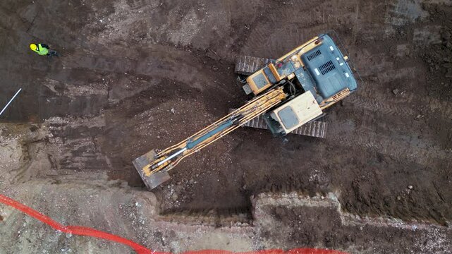 Ground excavator digging dirt and gravel at a construction site seen from above. Drone top down shot of industrial machinery excavating material during building of foundations for a modern stadium.
