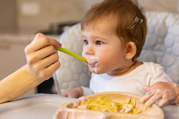 Mom feeding her baby girl with a spoon. Mother giving food to her eight-ten month child at home. Baby food. Children meal concept. High quality photo