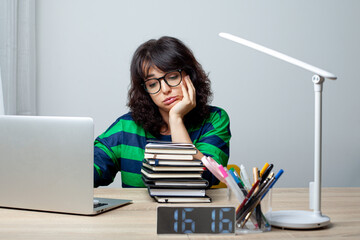 A woman in glasses and a striped green and navy sweater sits at a desk, looking bored and resting her head on her hand. She is surrounded by a laptop, a stack of books, pens, and a digital clock.
