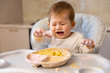Cute little toddler kid girl crying and eating while sitting in baby chair with messy face. Self-feeding concept. First solid food. Children meal concept. High quality photo