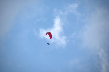 Paraglider with a red canopy soaring high in the sky under scattered clouds.