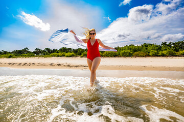Summer vacations. Beautiful mature woman in swimsuit holding scarf fluttering in wind walking in...