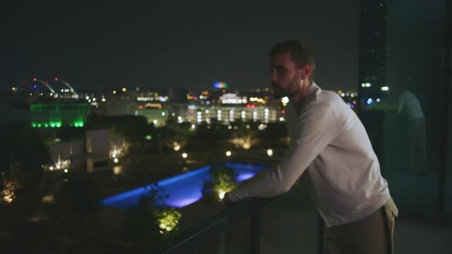 Serious man leans over the balcony with a view of a city at night