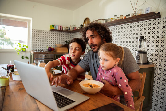 Father working on laptop with kids having breakfast in kitchen