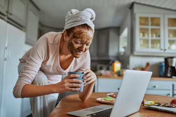 Woman with face mask working on laptop in kitchen during breakfast