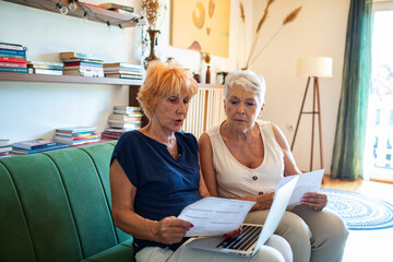 Naklejka premium Shocked senior lesbian couple holding bills looking at laptop on couch