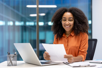 Confident young woman in modern office smiling at camera while holding document. Displays...