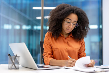 Young professional woman with curly hair and glasses working at desk, reviewing documents beside laptop, exuding focus and concentration in modern office