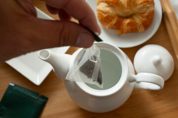 A view of a hand steeping a tea sachet into a ceramic teapot.