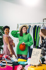 Cheerful young student friends having fun at home swap party, exchanging clothes, trying hats. Eco...