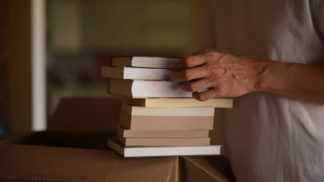 Closeup of man carefully packing books into cardboard box, preparing for move, organizing and looking forward to new beginning, close-up, slow motion. Concept of new life in new home relocation
