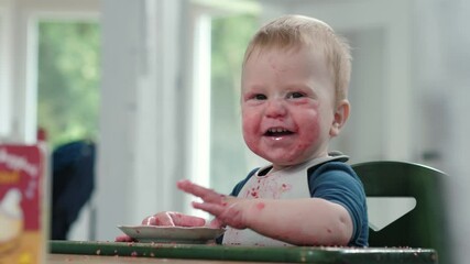 Dirty and cute baby with food stains sits happily in a high chair and playing with a plate, smiles looks at camera, clearly demonstrating the pure joy and inevitable mess that comes with eating