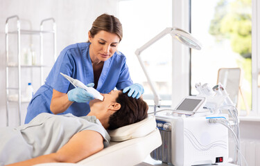 Obraz premium Focused young female cosmetologist performing facial skin cleansing with ultrasonic shovel on mature woman to enhance efficacy of skincare products and rejuvenate skin. Modern hardware cosmetology..