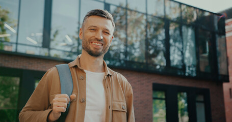 Satisfied Caucasian man glancing at camera while smiling with joy. Holding backpack on one shoulder. In background visible large building. Sun shining bright on attractive male. Happy person.