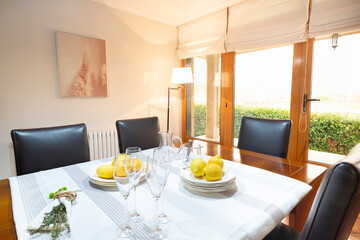 Natural wood dining table with five black leather chairs, dining room with French doors and white blinds, white tablecloth with gray stripes, champagne glasses, plates and lemons, chalet, country hous