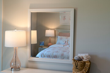 Cozy kids bedroom with a pastel-themed bed reflected in a white-framed mirror, featuring soft lighting, playful decor, and a woven basket with plush toys.