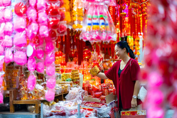 Happy Asian senior woman celebration Chinese lunar new year festival. Elderly retired woman in red blouse holding shopping bag walking and buying home decorative ornaments at Chinatown street market.