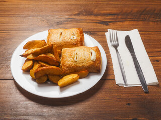 Dinner meal with golden wedges and two crispy fish and cheese pie on white plate and wooden table. Fine food with fried potatoes and staffed pastry. Simple basic dish.