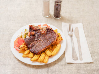 Dinner with premium t bone steak, fried potatoes chips with ridge, fresh salad with feta cheese on white plate and light color table cloth. Large portion of food with high quality products.