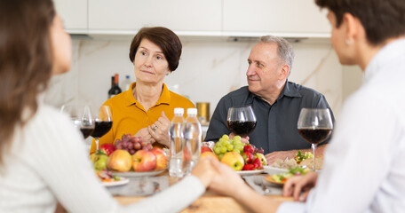 Friendly elderly parents enjoying family dinner with young adult son and daughter-in-law, sharing stories and laughing over wine and light snacks in cozy kitchen