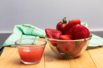 un vaso con jugo de frutas junto a un bol de vidrio lleno de fresas y frutillas sobre mesa de madera, al fondo un paño de cocina de tela verde claro con líneas verde oscuro desenfocado