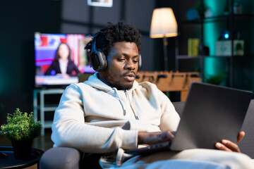 Cheerful man relaxing at home by watching TV series on laptop using headphones. Carefree person enjoying leisure time in RGB lit apartment at night, consuming entertainment media content