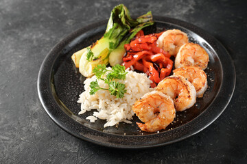 Fried prawns and vegetables such as bok choy and red bell peppers with rice, served on a black plate and dark slate, healthy homemade meal in Asian style, copy space
