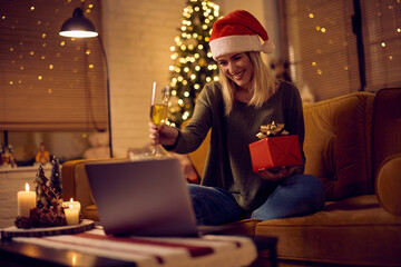 Happy woman holding present while toasting during video call on Christmas Eve.